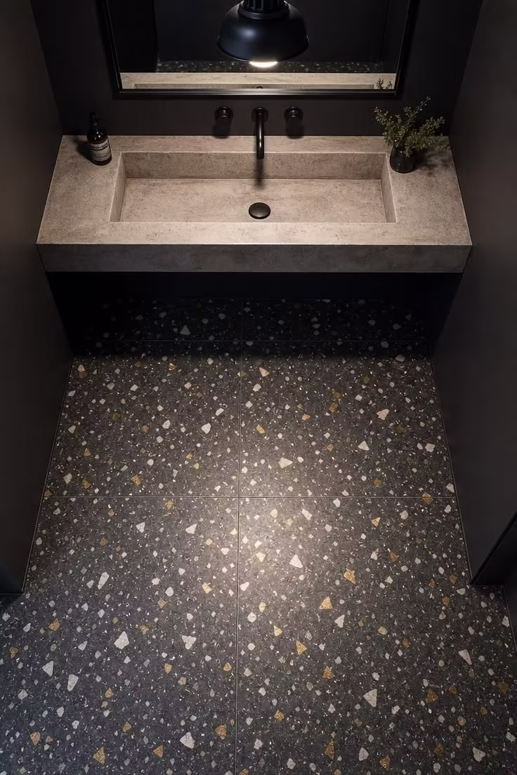 Dark terrazzo floor with brass and white chips beneath a concrete trough sink in a charcoal powder room