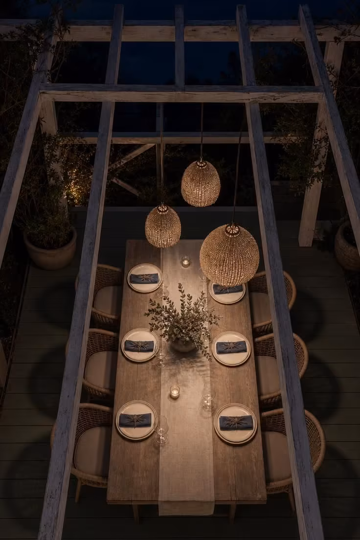 Overhead view of woven jute pendant lights hanging from whitewashed pergola rafters over a set dining table at dusk