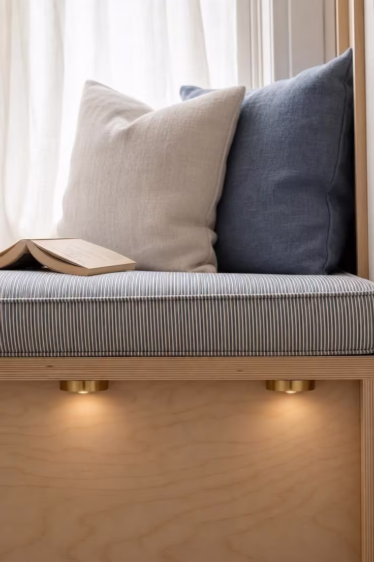 10 Cozy DIY Reading Nook Ideas You Can Build Today 7 Close-up of brass puck lights under a birch plywood window seat with striped ticking cushion and linen pillows