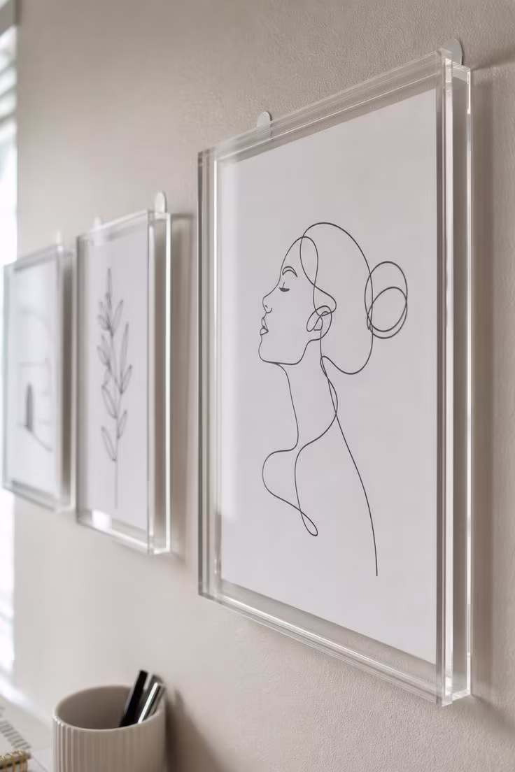 A close-up of a black-and-white minimalist line art print in a clear acrylic floating frame mounted on a dorm wall.