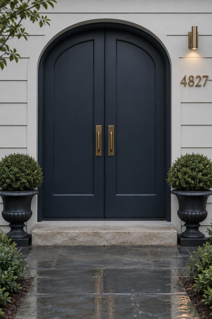 Arched double doors in deep blue-black with large brass house numbers on the pale wall beside the doorway and matching pulls on both leaves.