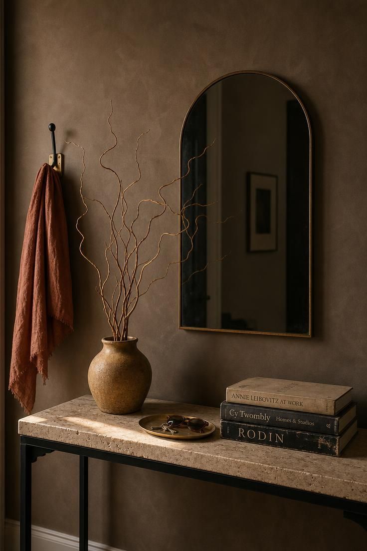 11 Inviting Eclectic Entryway Ideas for an Artful Home 1 Narrow foyer wall with travertine console, arched smoked-glass mirror, dried curly willow branch, and stacked art books in warm golden light.