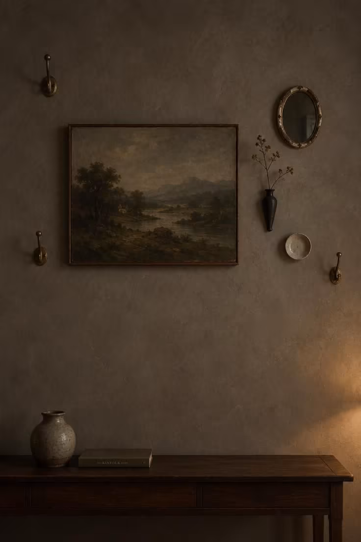 13 Warm Eclectic Farmhouse Decor Ideas for a Collected Home 7 Large weathered landscape painting hung low on a plaster wall with small brass hooks and a dried branch nearby.