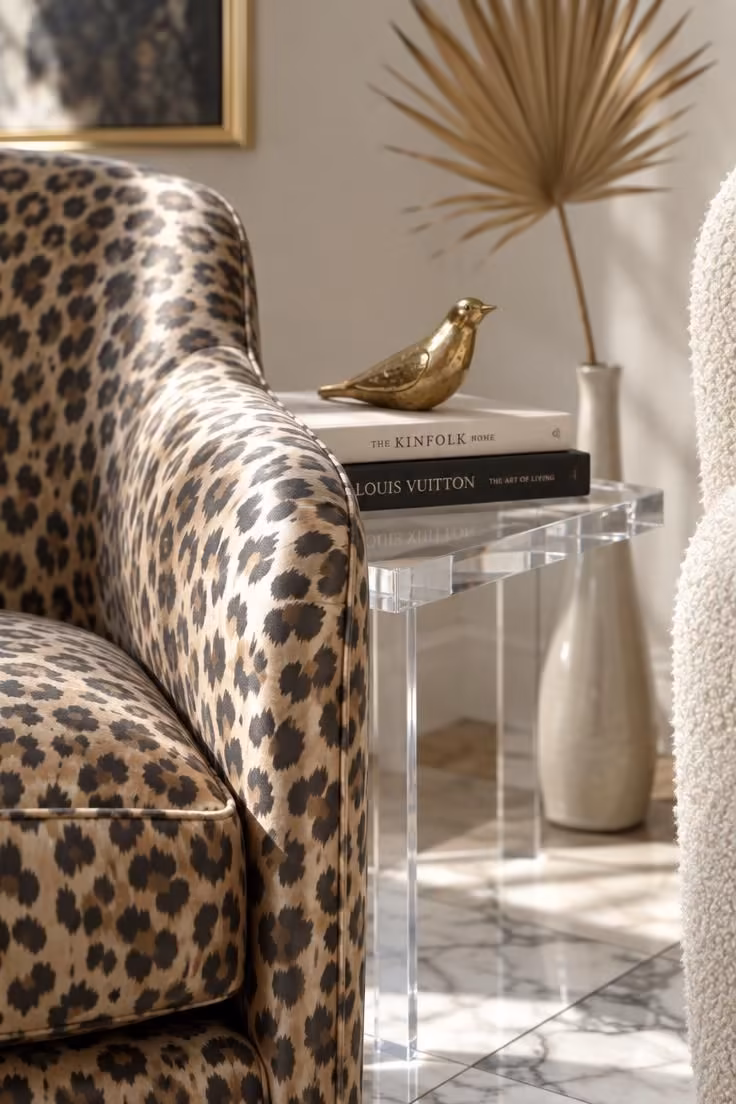13 Warm Eclectic Glam Decor Ideas That Feel Collected 7 Detail of a leopard-print silk-blend accent chair arm beside a clear acrylic side table with books and a small brass object.