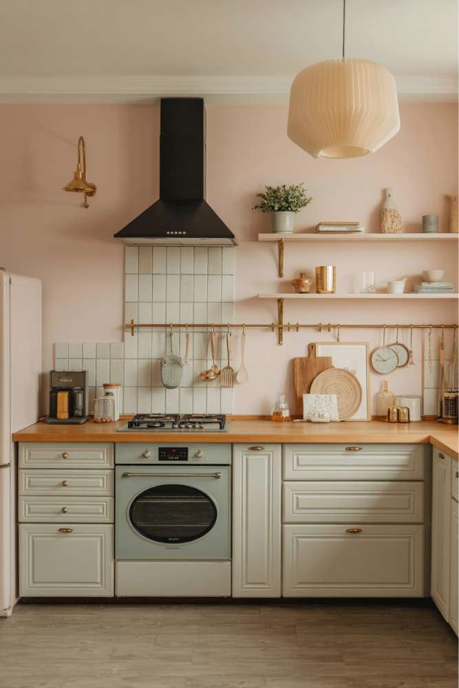 32 Eye-Catching Eclectic Kitchen Design Ideas to Love 11 Modern kitchen with pale pink walls, sage green cabinets, wooden countertop, brass hardware, beige paper lantern pendant, and minimalist design