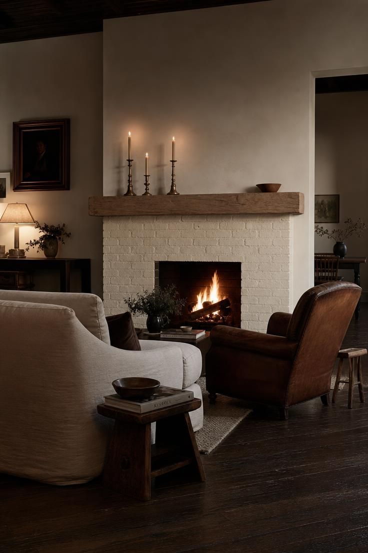13 Eclectic Mid Century Modern Ideas for Warm Lived-In Rooms 8 A warm white brick fireplace with an unfinished oak mantel, mismatched brass candlesticks, low sofa, and worn leather chair in a moody room with dark hardwood floor.