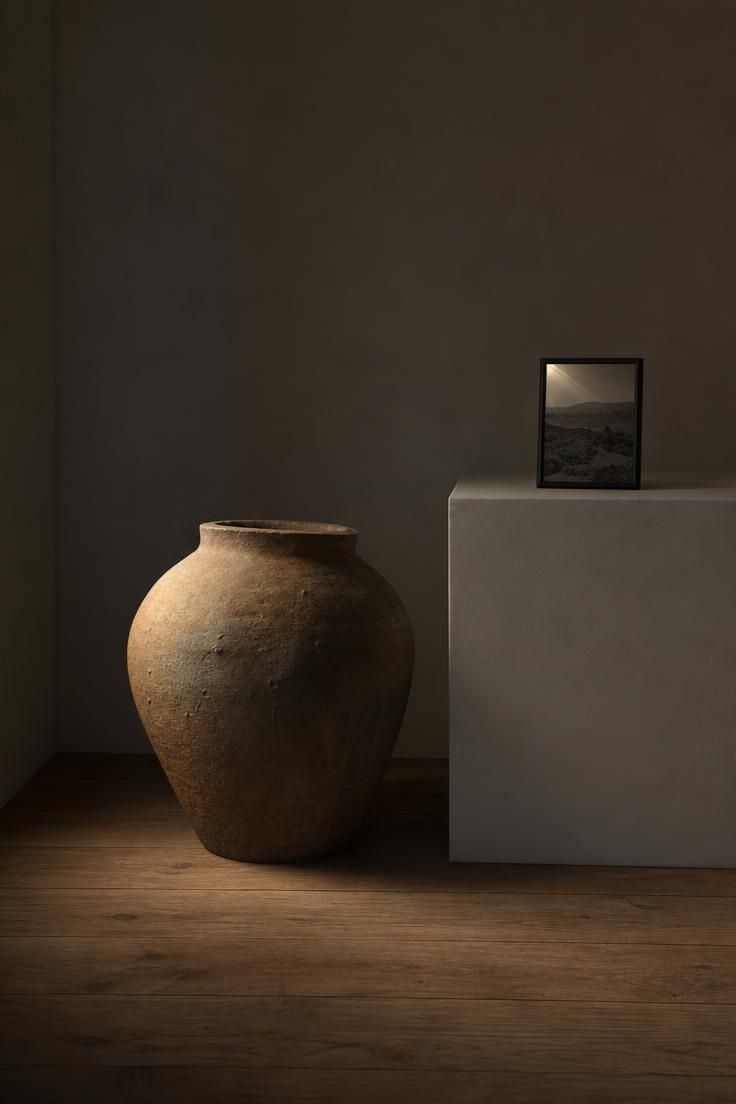 Large unglazed stoneware vessel on floor beside white plaster plinth with small black-framed photograph in moody low light.