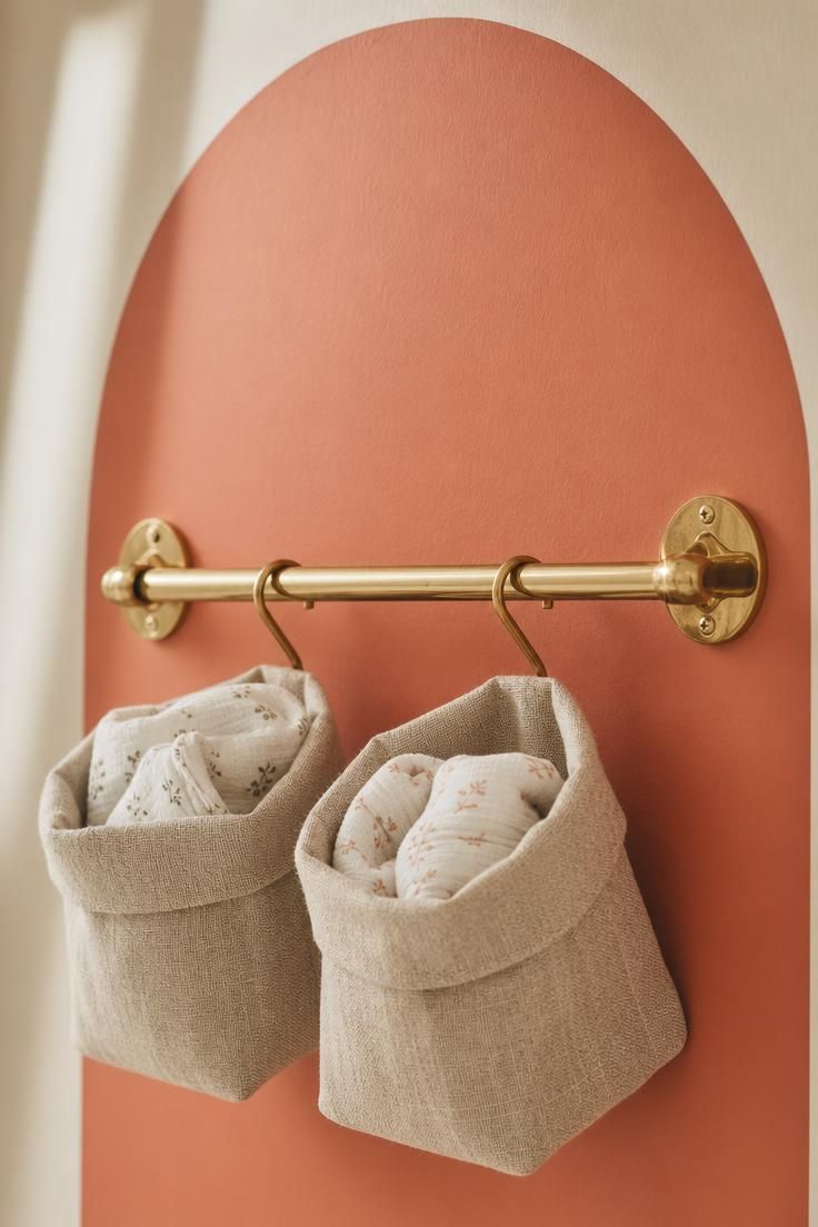 11 Cozy Eclectic Nursery Ideas for a Calm, Happy Baby 7 Wall-mounted brass rail with S-hooks holding two natural linen storage baskets above a single soft coral painted arch in a boho nursery.