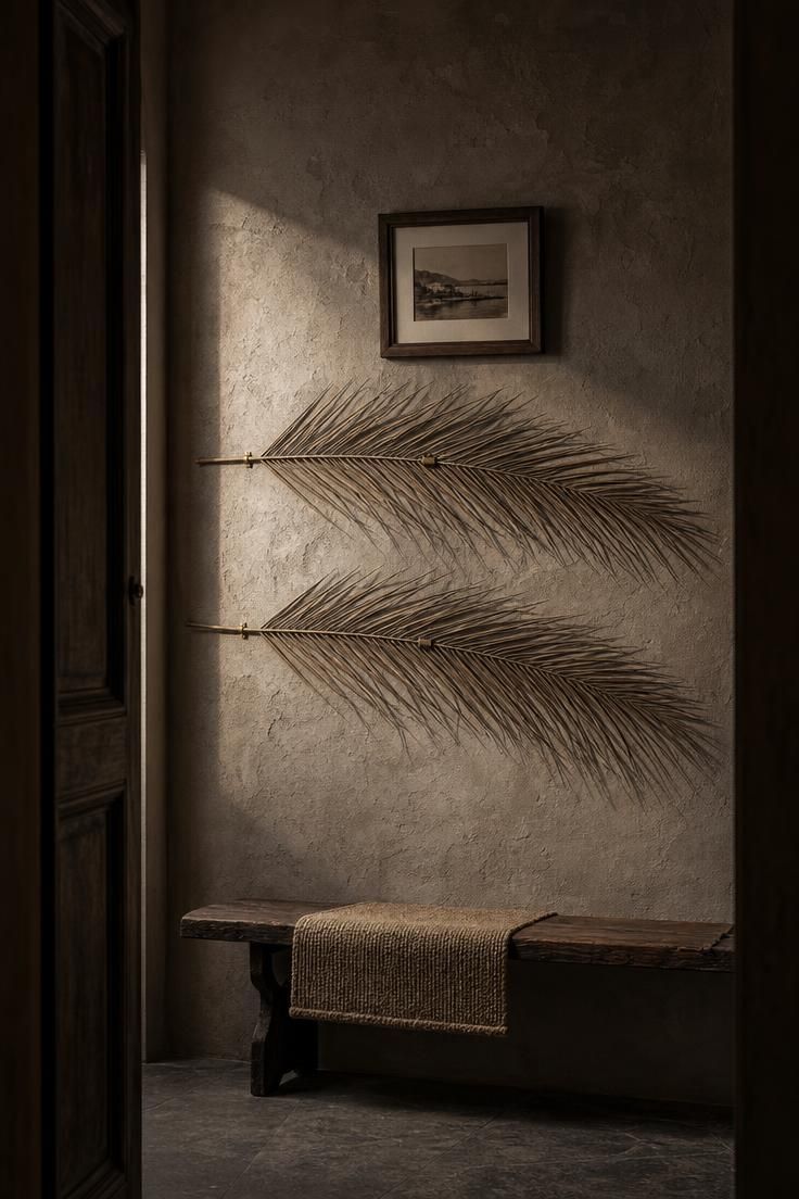 11 Warm Eclectic Wall Decor Ideas You Will Love Now 4 Two dried palm fronds mounted across a dark taupe plaster hallway wall, with a small framed photograph above and a low wooden bench below.