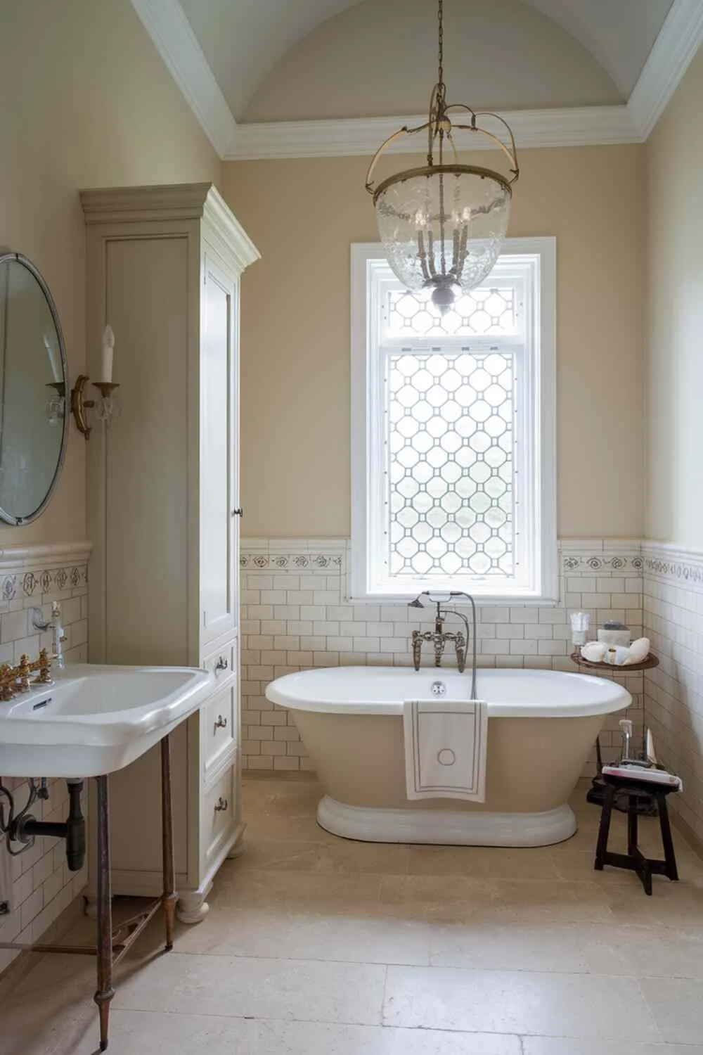 21 Elegant Bathroom Design Ideas for a Luxurious Look 13 Classic bathroom with centered freestanding bathtub surrounded by beige and white tiles, ornate chandelier, vintage-style sink with brass faucet, tall white cabinet, beige tiled floor, high ceiling, and decorative window