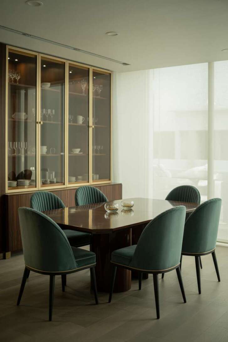 Elegant dining room with dark wood rectangular table, deep teal velvet chairs with gold frames, and glass-front built-in cabinets