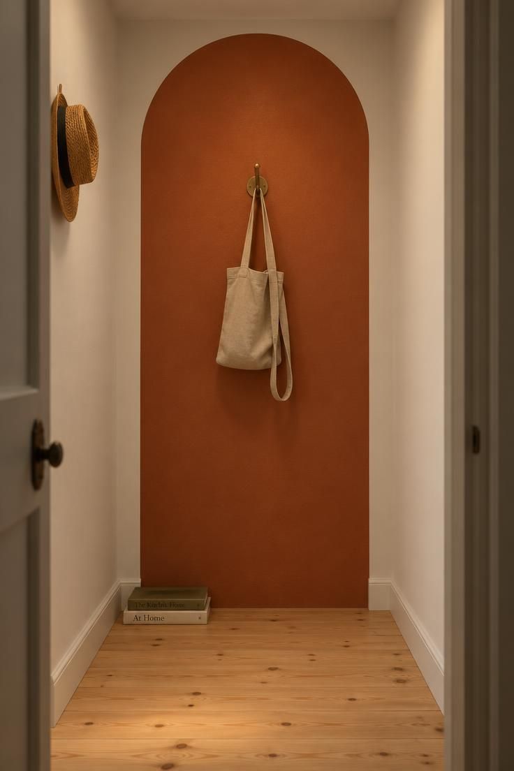 A terracotta painted arch on a white hallway end wall with a brass hook and canvas tote bag, seen from a doorway in moody low light with light pine flooring.