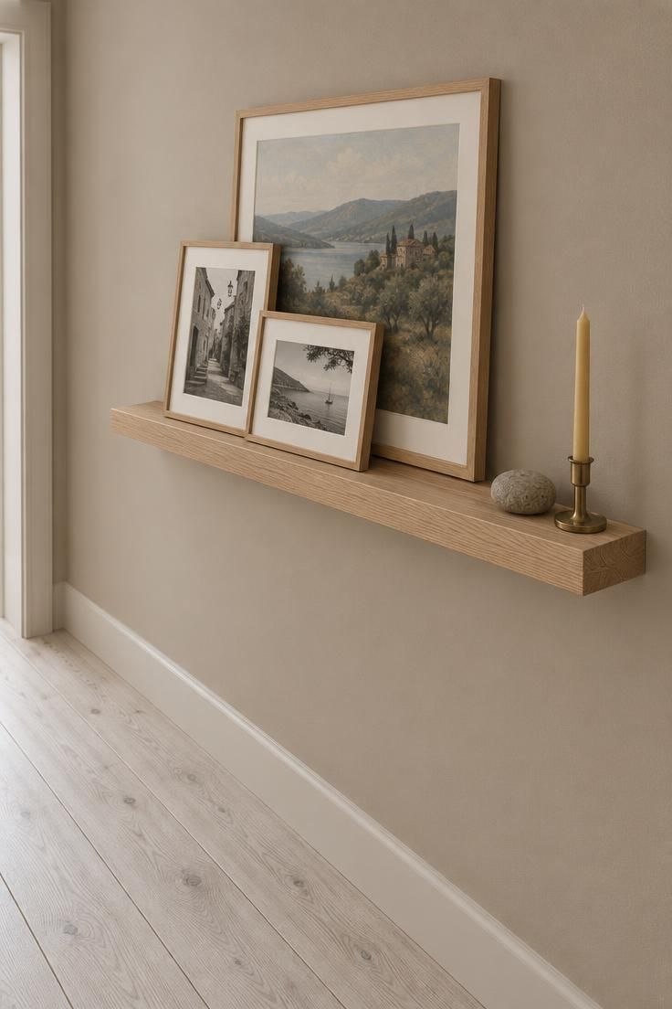 A floating oak picture ledge holding three layered framed prints at the end of a hallway with a soft clay wall and whitewashed wood flooring under diffused light.