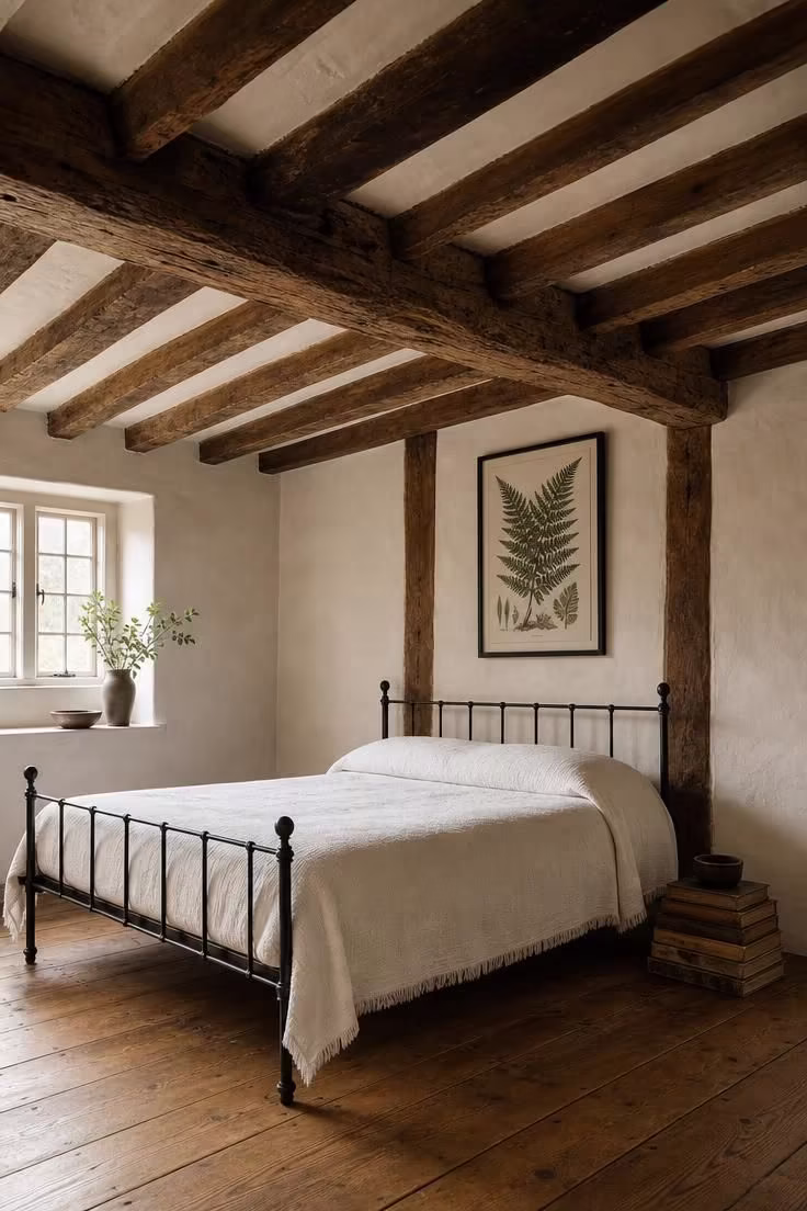 14 Cozy English Cottage Bedroom Ideas Full of Character 2 Exposed oak ceiling beams above a low black iron bedstead with a white coverlet and a framed fern print in an english cottage bedroom.