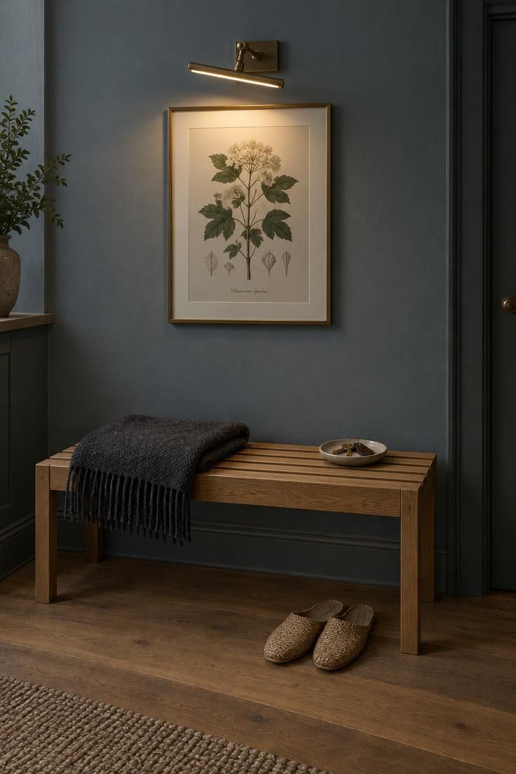 Oak slat bench with wool throw below a framed botanical print lit by a brass picture light, on a blue-gray wall with woven slippers on the floor.