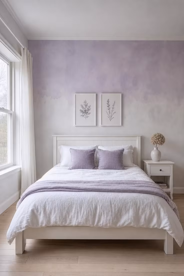 Hand-painted watercolor accent wall in lavender and dove behind a white bed with pale lilac throw in cool overcast light