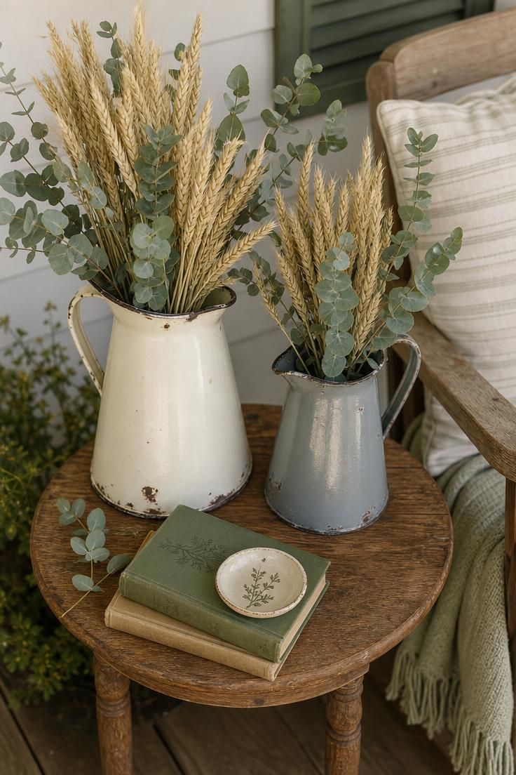 17 Cozy Fall Outdoor Decor Ideas for a Warm Welcome 14 Two chipped enamelware pitchers holding dried wheat and eucalyptus on a wood side table in soft diffused light.