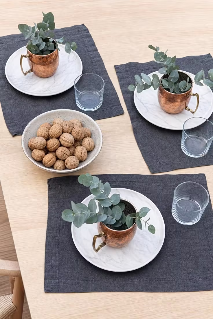 15 Fall Table Decor Ideas That Make Guests Want to Linger 8 Copper mugs holding eucalyptus stems at each place setting on white plates with charcoal placemats on a light birch dining table.