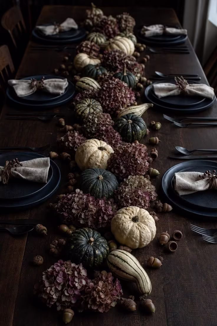 15 Fall Table Decor Ideas That Make Guests Want to Linger 14 A harvest runner of mini gourds acorns and dried hydrangeas stretching down a dark farmhouse table with deep blue plates in moody low light.