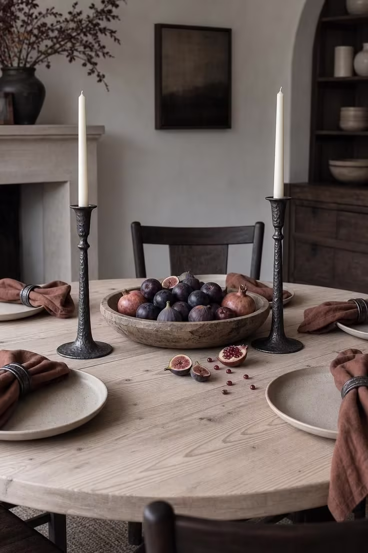 15 Fall Table Decor Ideas That Make Guests Want to Linger 15 Iron candlestick holders flanking a wooden bowl of plums figs and pomegranates on a pale pine table under cool overcast light.