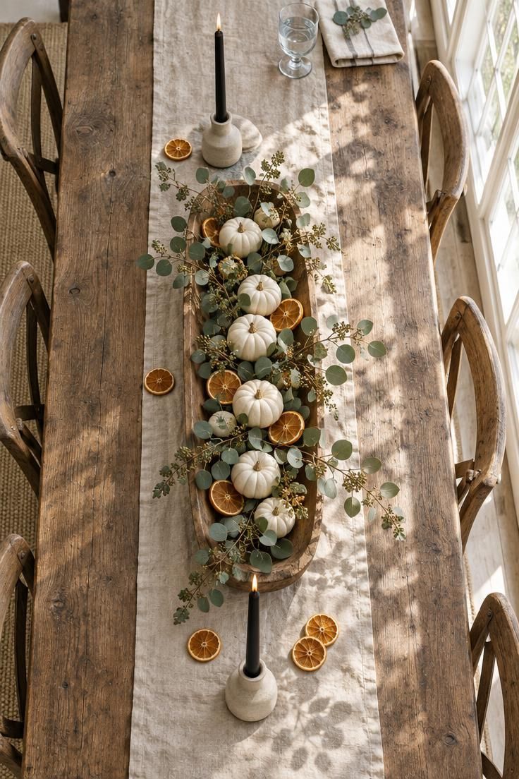 An overhead view of a linen runner and raw wood dough bowl filled with mini white pumpkins, dried orange slices, and eucalyptus, with black taper candles in stoneware holders at each end.
