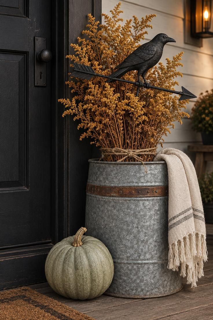 11 Whimsical Farmhouse Halloween Porch Decor Ideas 4 A black iron crow weathervane lying across dried goldenrod stems in a galvanized barrel next to a sage-green pumpkin on a porch.