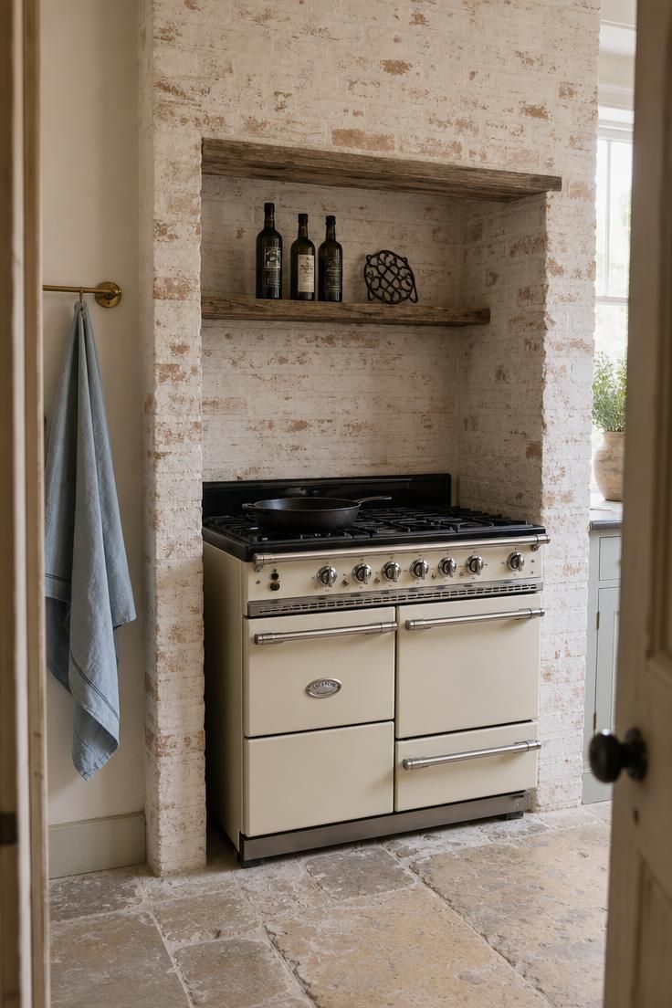 Cream enamel range cooker set inside a whitewashed brick alcove with a reclaimed wood shelf above in a french cottage kitchen with soft diffused light.