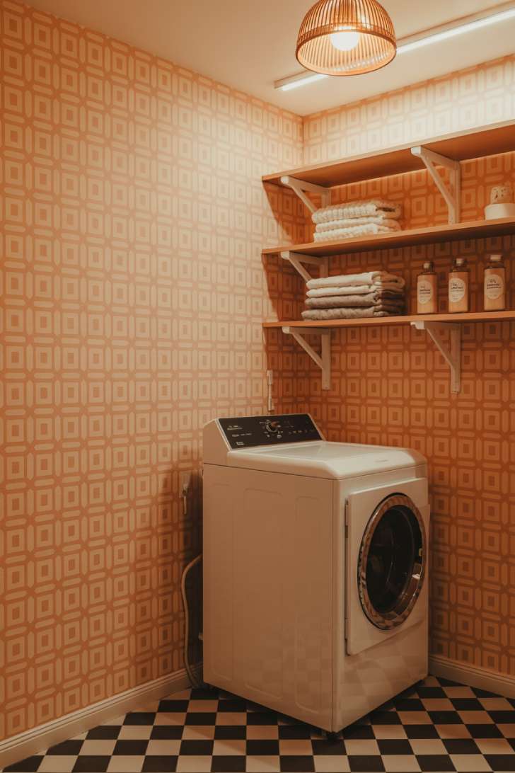 A laundry room wrapped in coral and cream geometric retro wallpaper with open wooden shelving, checkered floors, and a woven pendant light