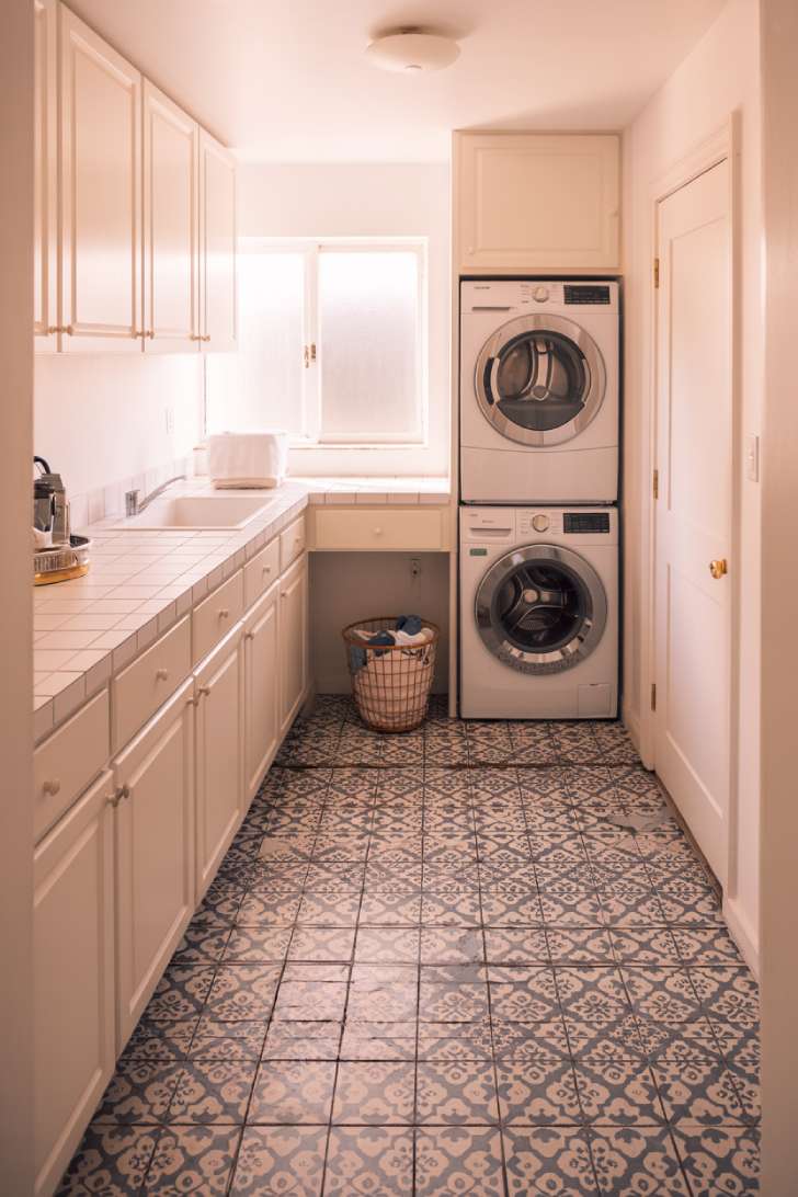 An elevated-angle shot of a laundry room with navy and white Moroccan patterned floor tiles, white cabinets, and a stacked washer and dryer
