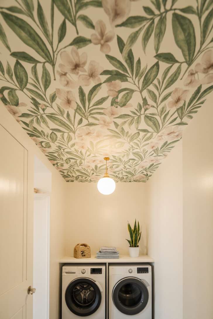 An upward-angle shot of a small laundry room with botanical wallpaper on the ceiling, white walls, a stacked washer/dryer, and a white globe pendant