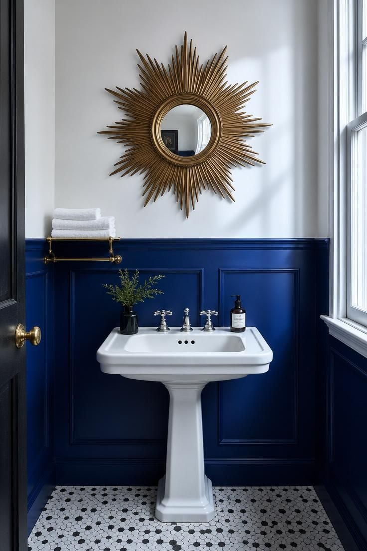 Powder room with cobalt blue wainscoting on lower walls, white upper walls, and a large vintage brass starburst mirror above a white pedestal sink in cool overcast light.