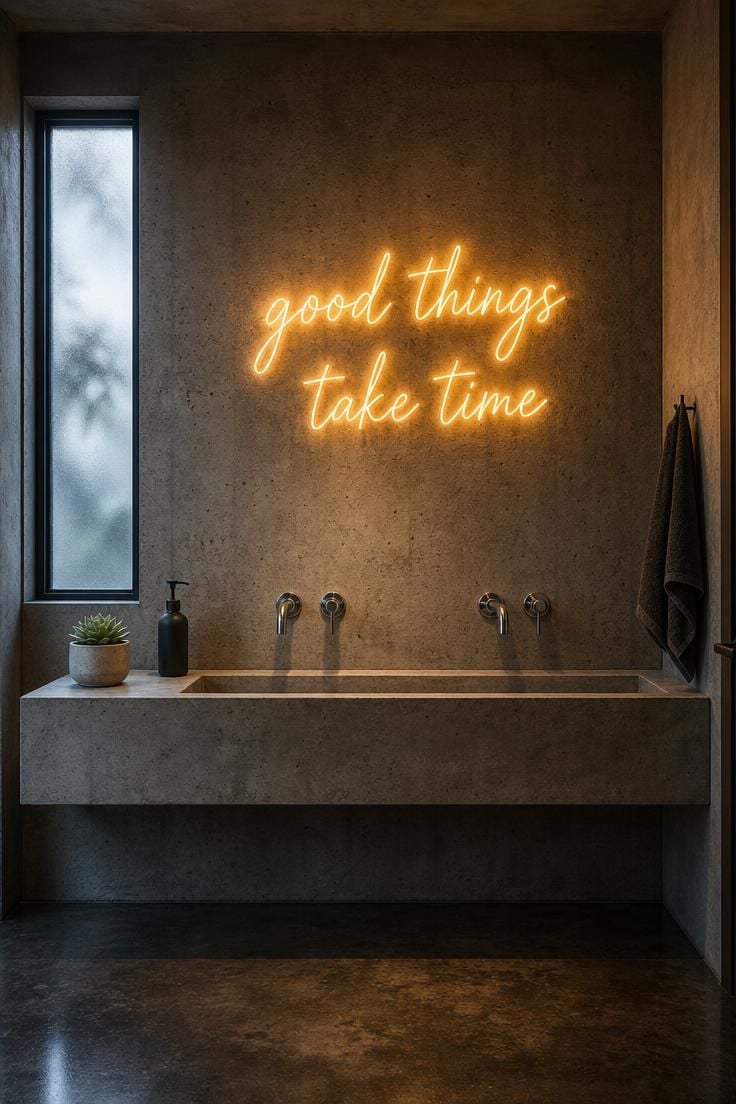 Raw concrete powder room wall with a warm amber neon sign above a concrete trough sink, cool overcast light from a frosted window mixing with the neon glow.