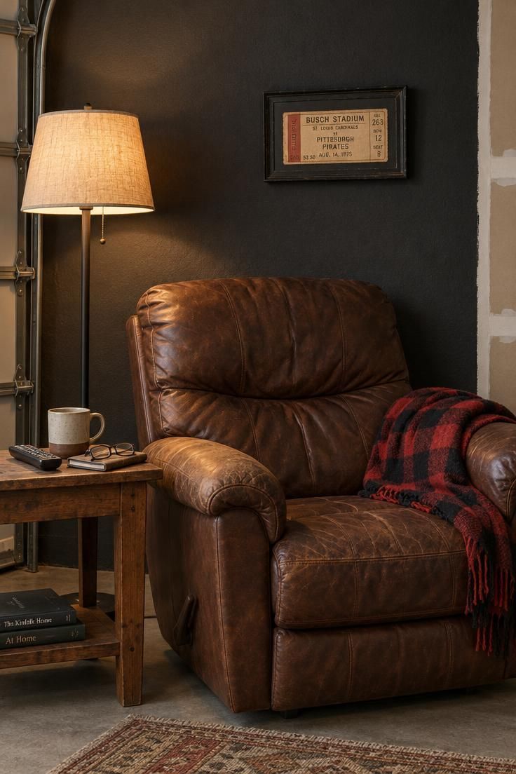 A charcoal-black matte accent wall in a garage behind a worn brown leather recliner, with a small side table, a plaid blanket, and a thrifted framed ticket above.