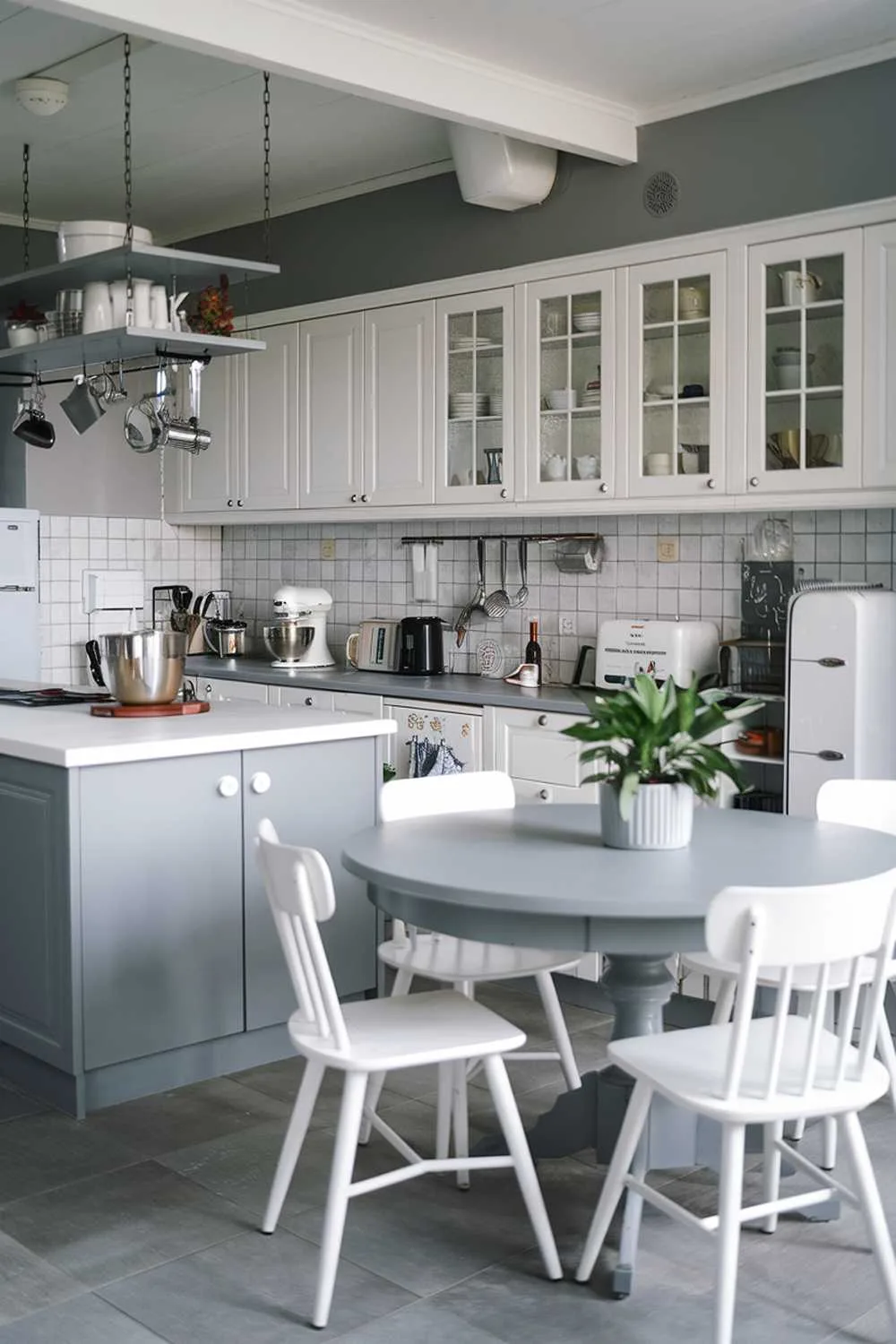 21 Inspiring Gray and White Kitchen Ideas for a Modern Look 6 A gray and white kitchen decor. The kitchen has a gray island with a white countertop and a gray, round dining table with white chairs. There are white cabinets and a gray backsplash. A potted plant sits on the counter. There are various kitchen appliances, including a mixer, a toaster, and a coffee maker. A few utensils are hung above the island. The floor is made of gray tiles.
