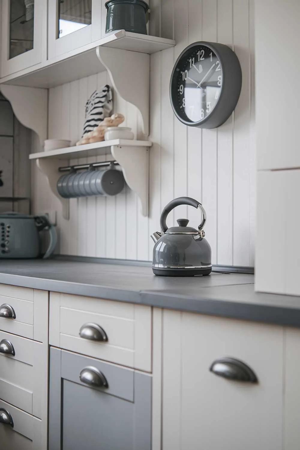 21 Inspiring Gray and White Kitchen Ideas for a Modern Look 9 A close-up shot of a cozy gray and white kitchen decor. There's a white cabinet with gray drawers and a gray counter on the bottom. There's a gray kettle on the counter. On the wall above the counter, there's a gray clock and a few white shelves with gray handles.