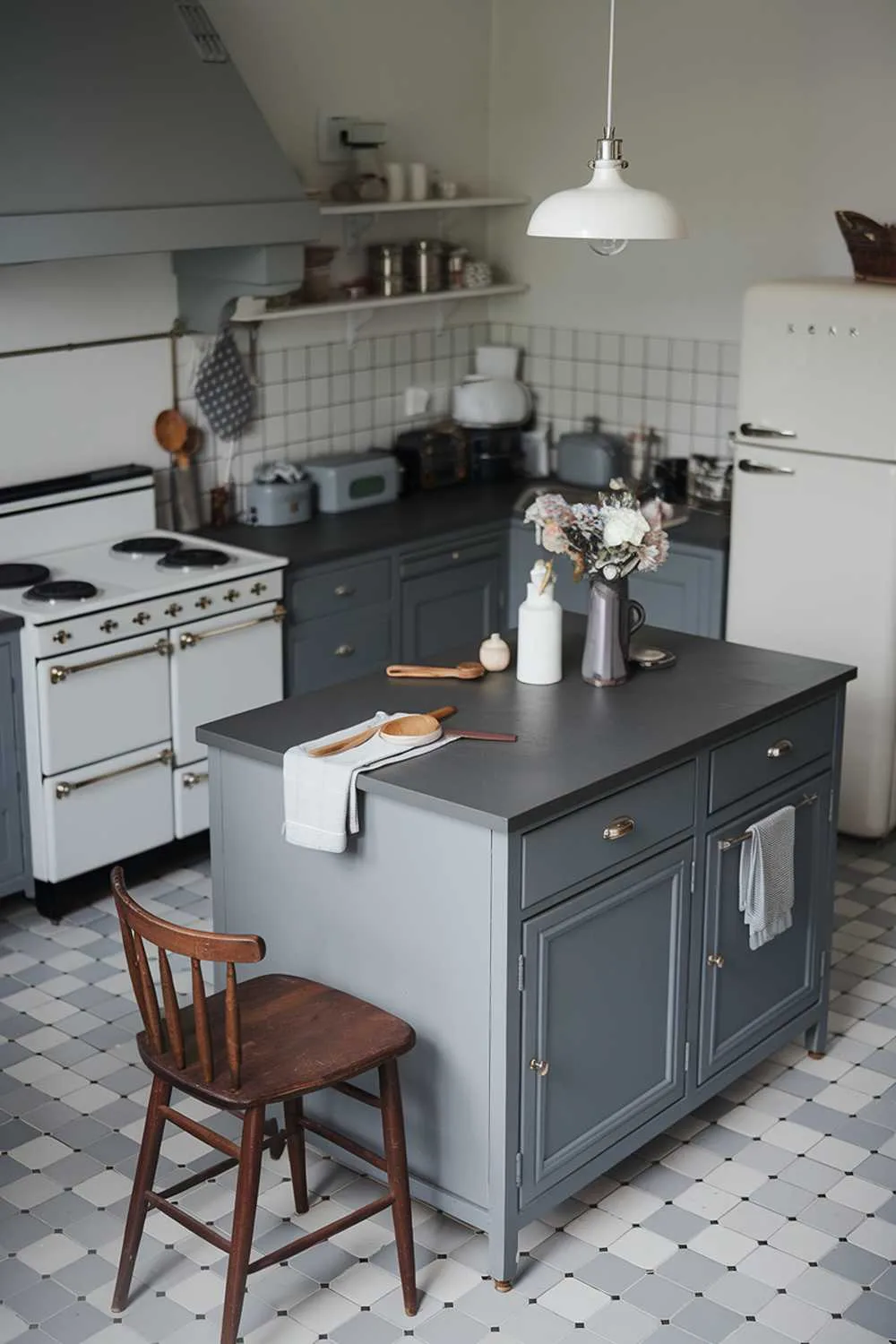 21 Inspiring Gray and White Kitchen Ideas for a Modern Look 10 A cozy, modern gray and white kitchen with a gray island in the center. The island has a dark gray countertop and a few items, including a vase of flowers and a wooden spoon. There is a white stove with four burners on the left wall. Above the stove, there is a gray hood. On the right wall, there is a white refrigerator. The floor is gray and white tiles. A wooden chair is placed near the island. A white pendant light hangs over the island.