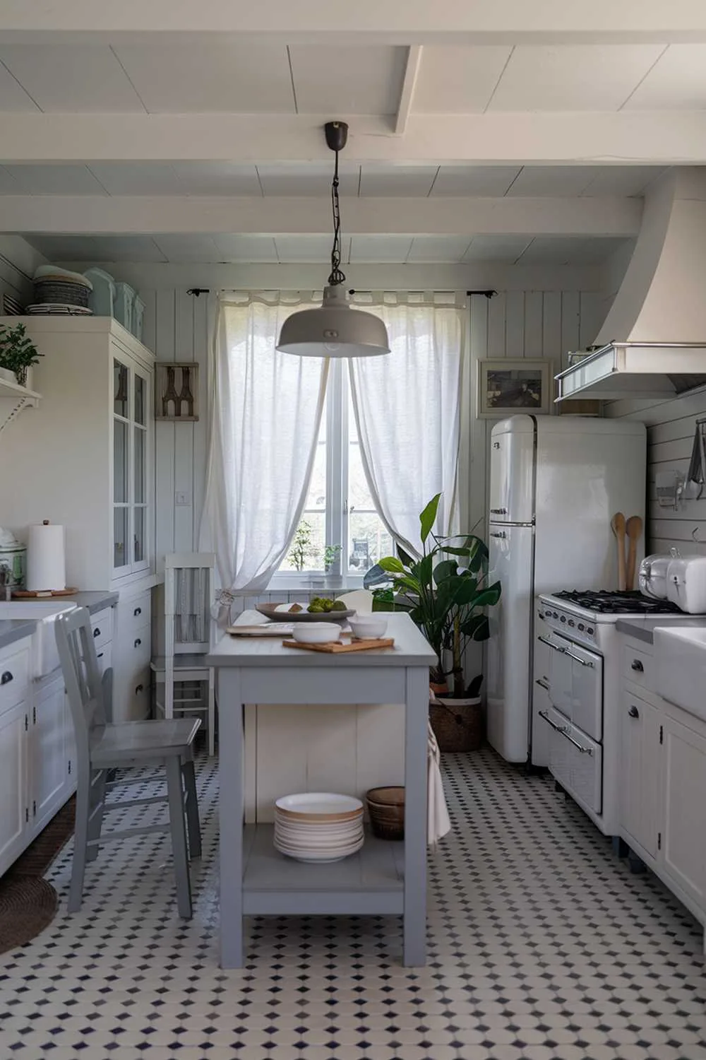 21 Inspiring Gray and White Kitchen Ideas for a Modern Look 11 A cozy white kitchen with gray touches, featuring a center island with a gray countertop and a gray pendant light. There's a white cabinet, a white refrigerator, and a white range hood over the stove. The floor is covered with gray and white checkered tiles. A gray chair is placed near the island. There's a potted plant near the refrigerator. The room has a window with white curtains. The room has a wooden spoon and a white plate on the counter.