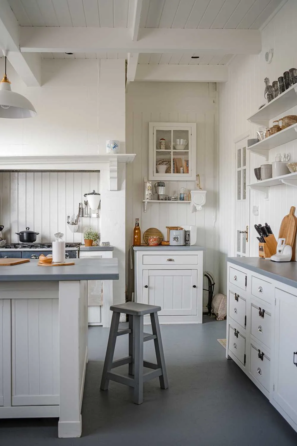 21 Inspiring Gray and White Kitchen Ideas for a Modern Look 13 A white kitchen with gray touches. The kitchen has a white island with a gray countertop and a white cabinet above. There is a gray stool near the island. The walls are white and have gray accents. There is a white cabinet near the wall with a gray countertop. The floor is gray. There is a white pendant light hanging over the island.