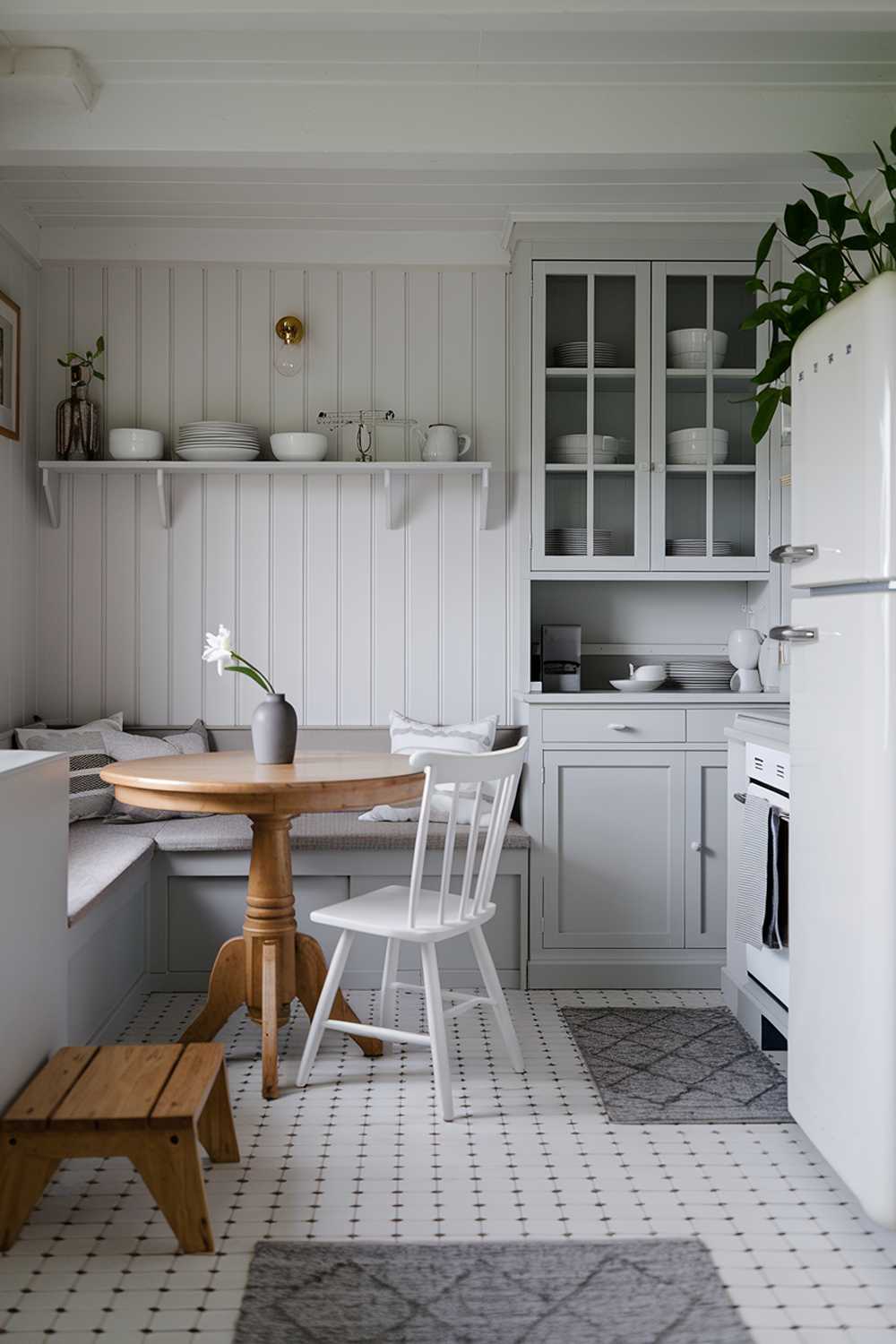 A cozy white kitchen with light gray touches. The room has a breakfast nook with a round wooden table and white chairs. A gray bench sits along the wall. There's a white cabinet with a glass door, holding some white and gray dishes. A gray vase with a single white flower sits on the counter. There's a white refrigerator with a gray handle. A potted plant stands next to the refrigerator. The floor is covered with white tiles. The room has a wooden step stool and a gray rug. The walls have a white shelf and a simple light fixture.