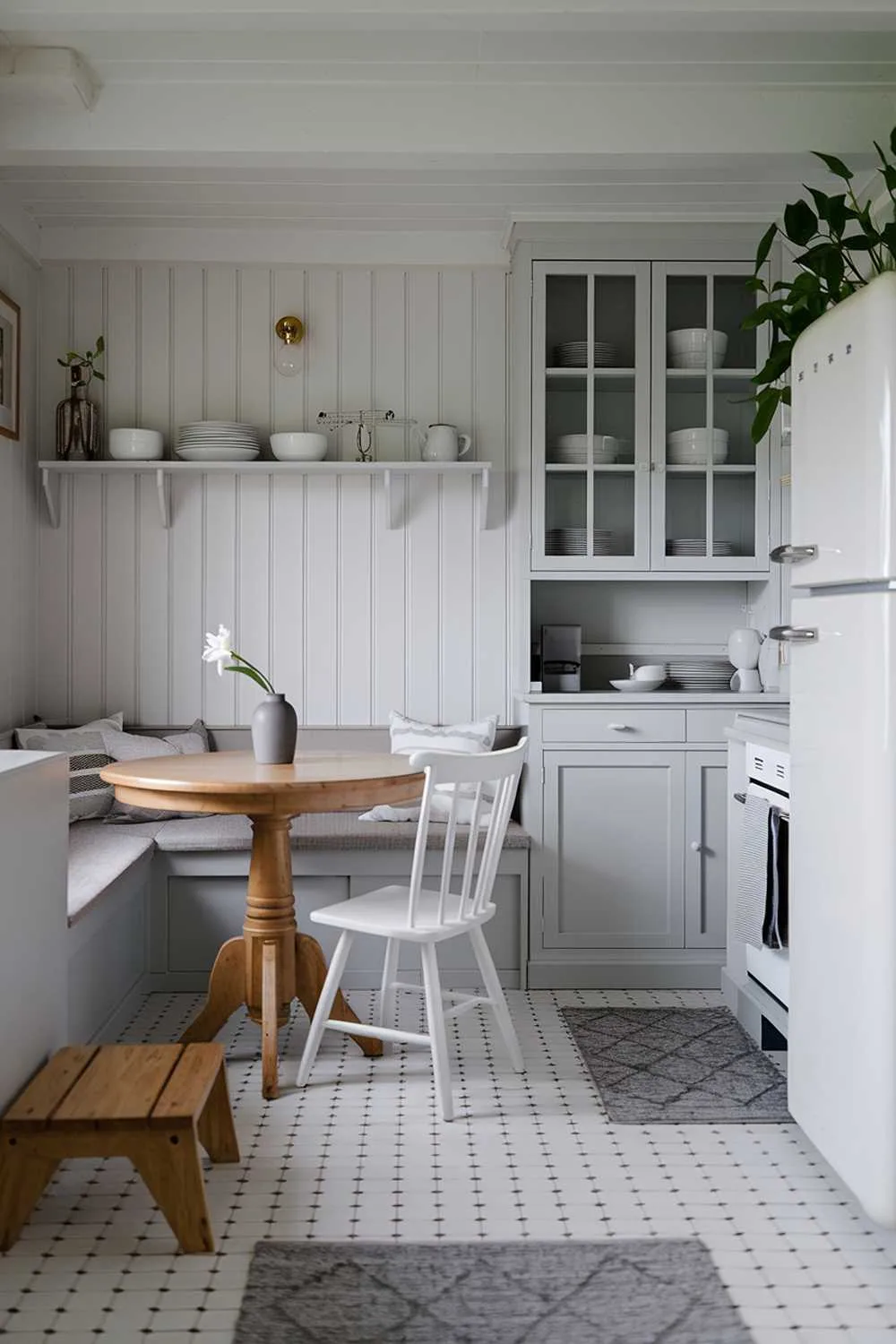 21 Inspiring Gray and White Kitchen Ideas for a Modern Look 16 A cozy white kitchen with light gray touches. The room has a breakfast nook with a round wooden table and white chairs. A gray bench sits along the wall. There's a white cabinet with a glass door, holding some white and gray dishes. A gray vase with a single white flower sits on the counter. There's a white refrigerator with a gray handle. A potted plant stands next to the refrigerator. The floor is covered with white tiles. The room has a wooden step stool and a gray rug. The walls have a white shelf and a simple light fixture.