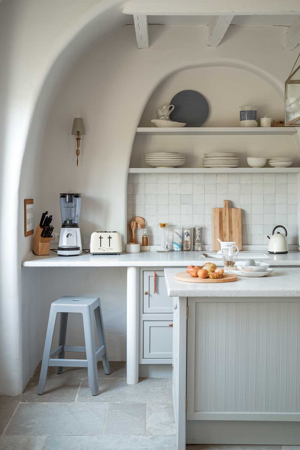 A white kitchen with light gray touches. The kitchen has a smoothie maker, a toaster, and a coffee maker on a white countertop. There is a gray stool near the counter. The walls and cabinets are white, and there is a light gray kitchen island with a few items on it. The floor is made of light gray tiles.