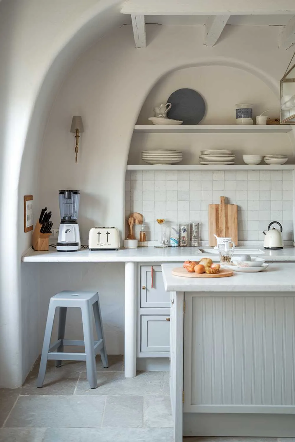 21 Inspiring Gray and White Kitchen Ideas for a Modern Look 17 A white kitchen with light gray touches. The kitchen has a smoothie maker, a toaster, and a coffee maker on a white countertop. There is a gray stool near the counter. The walls and cabinets are white, and there is a light gray kitchen island with a few items on it. The floor is made of light gray tiles.
