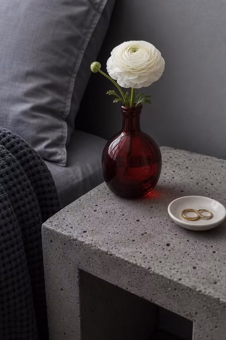 Ruby red glass bud vase with white ranunculus on a concrete nightstand in cool overcast light
