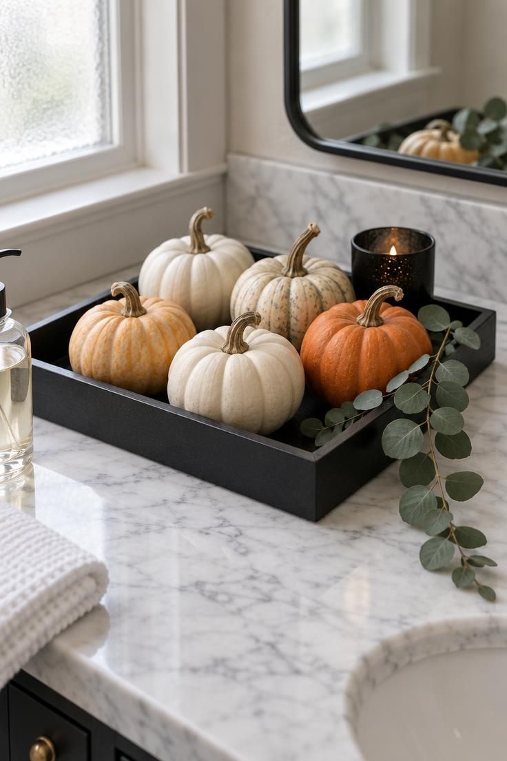 Matte black tray on a white marble vanity holding five miniature pumpkins in cream and light orange with a trailing eucalyptus sprig and a small black votive holder.