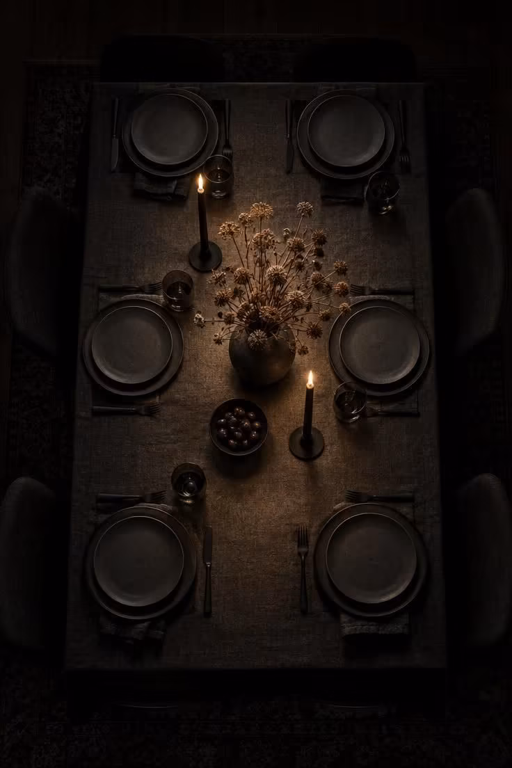 11 Hauntingly Easy Halloween Party Decor Ideas for Home 5 Overhead view of a dining table lit by a single warm pool of light, with matte charcoal plates and low dark centerpiece fading into shadow at the edges.