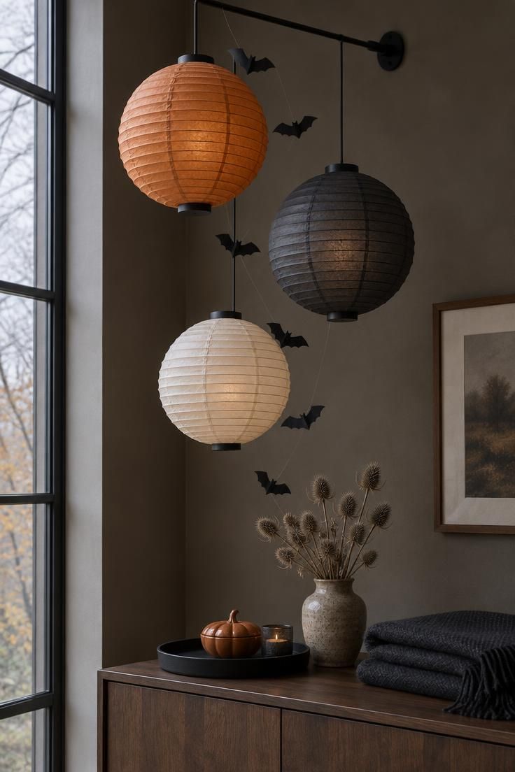 Three paper lanterns in muted orange, black, and cream hanging at different heights from a black rod with paper bats between them