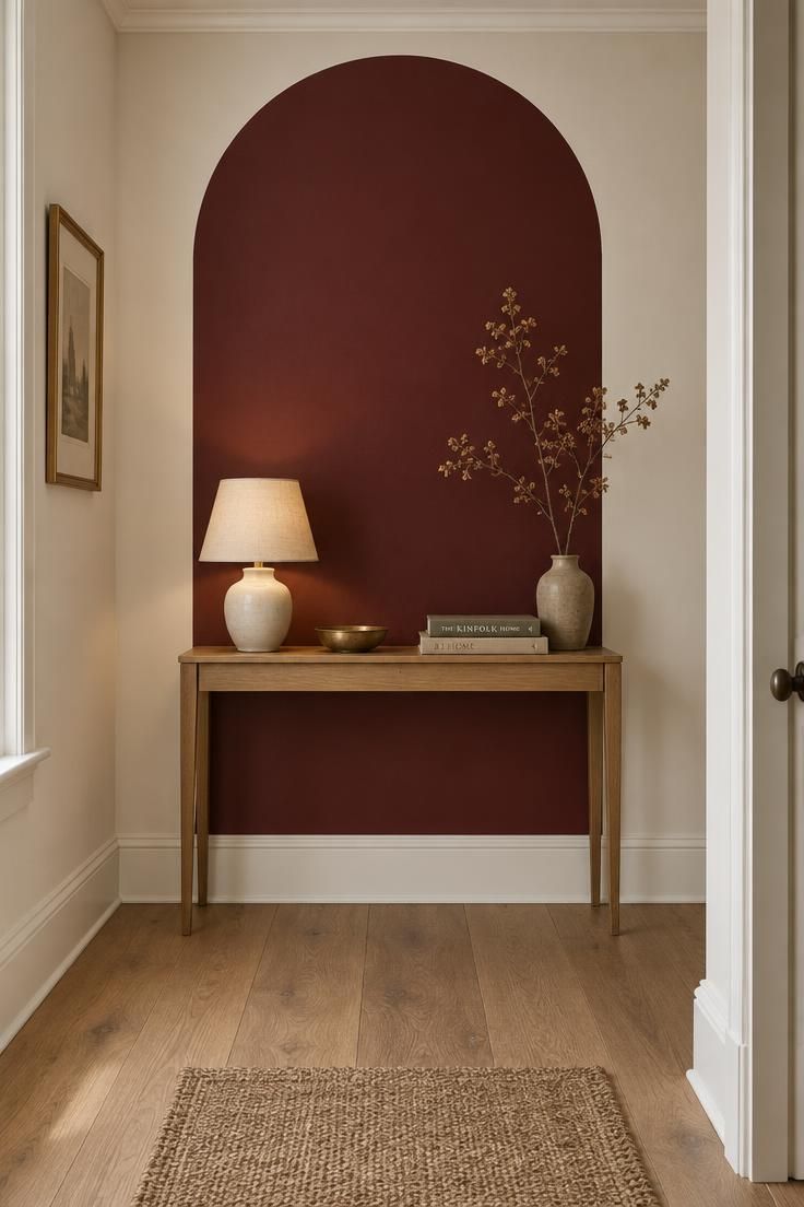 A hallway end wall with a tall oxblood painted arch behind a slim oak console holding a small ceramic lamp and brass dish, surrounded by warm white walls.