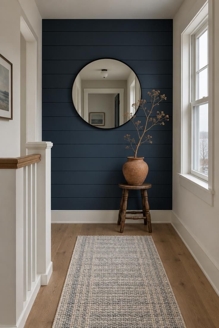 A landing end wall in deep inky blue horizontal shiplap with a round black-framed mirror centered, a small stool with a terracotta pot, and cool daylight catching the seam ledges.