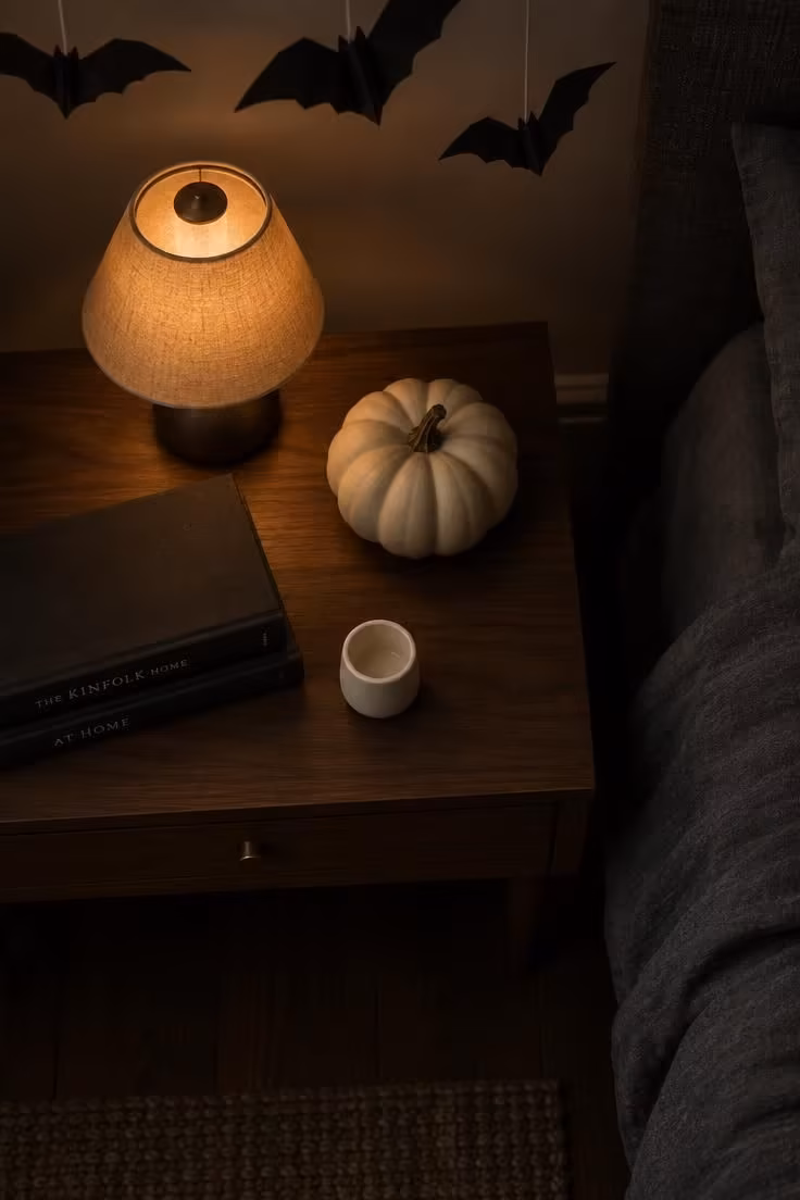 13 Indoor Halloween Decor Ideas for a Cozy October Home 12 Overhead view of a nightstand with a cream pumpkin, matte black book stack, small votive, and suspended paper bats above.
