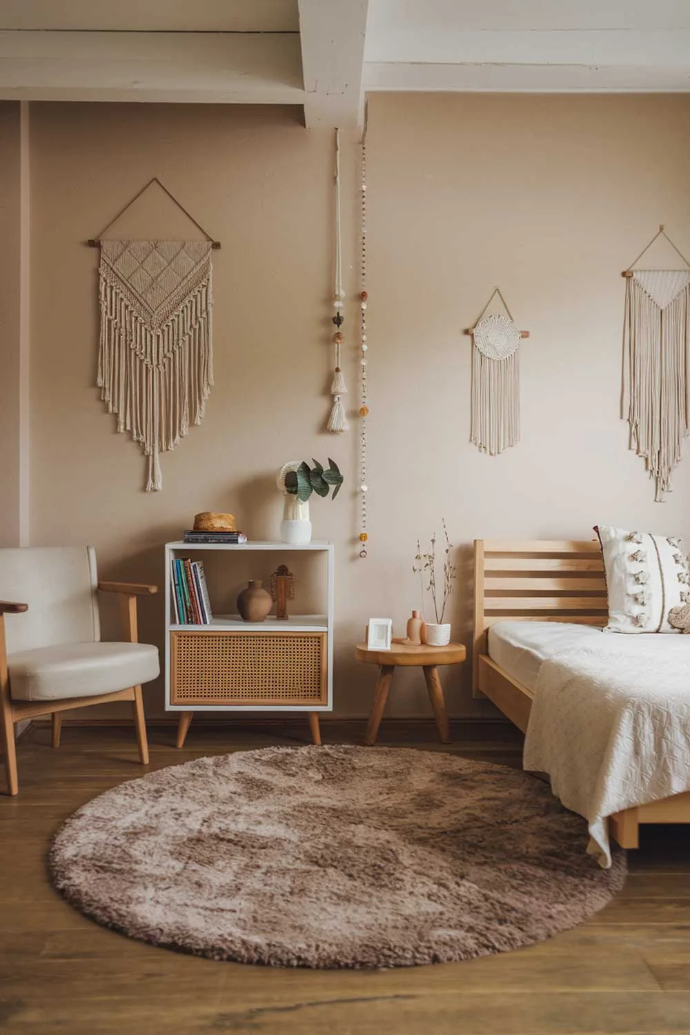 A cozy japandi bedroom interior design. The room has a wooden floor and is furnished with a plush beige rug, a wooden bed frame with a white mattress, and a few pieces of wooden furniture. There is a white bookshelf with a few books and decorative items. A beige chair with a wooden frame is placed near the bed. The walls are painted in soft beige and have a few hanging decorations.